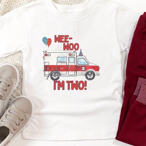 May include: White t-shirt with a red and white ambulance graphic, two balloons, and the text "Wee-Woo I'm Two!"