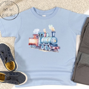 May include: A light blue t-shirt featuring a watercolor illustration of a train. The train is in shades of blue, red, and orange, with smoke billowing from the smokestack. The text "SIZES 6M-6T AVAILABLE" is in a white circle. Gray pants and blue shoes are also in the image.