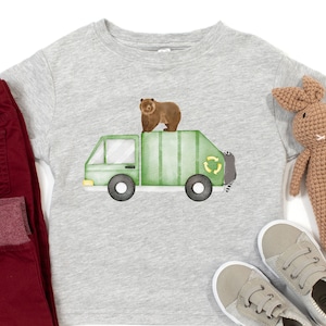 Puede incluir: Una camiseta gris con un camión de basura verde y un oso marrón en la parte superior. El camión tiene un símbolo de reciclaje en el lateral.