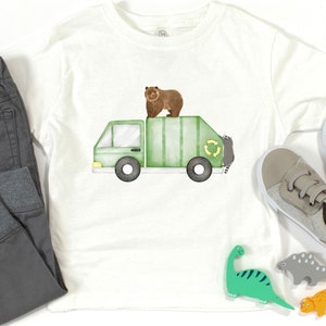 May include: White t-shirt featuring a watercolor illustration of a green garbage truck with a brown bear on top. The truck has black wheels and a recycling symbol. Gray pants and shoes are next to the shirt.