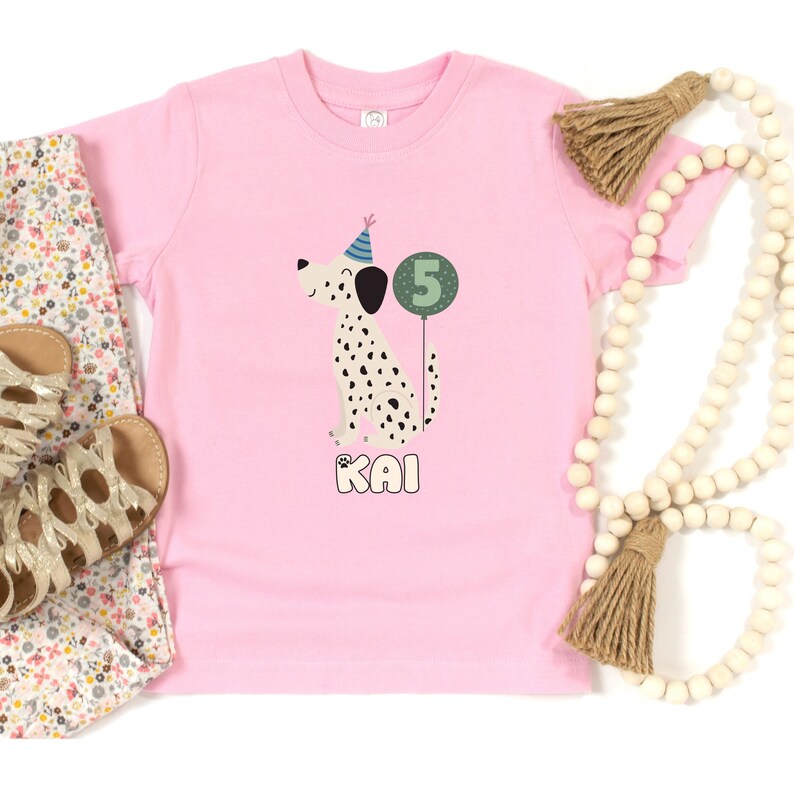 May include: A pink toddler t-shirt featuring a Dalmatian dog wearing a party hat and holding a number 5 balloon. The name "KAI" is printed below the dog. The shirt is paired with floral pants and gold sandals.