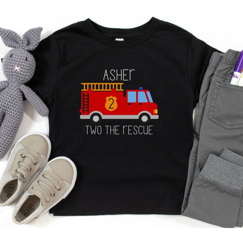 Puede incluir: Una camiseta negra de manga corta con el gr&aacute;fico de un cami&oacute;n de bomberos rojo, una escalera amarilla y un escudo con el n&uacute;mero 2. El texto ASHER est&aacute; impreso sobre el cami&oacute;n y el texto TWO THE RESCUE est&aacute; impreso debajo del cami&oacute;n.