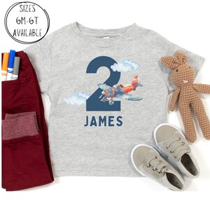 May include: A light gray toddler t-shirt with a navy blue number 2 and the name "JAMES" printed on the front. A watercolor illustration of a red and blue airplane and clouds is next to the number. Sizes 6M-6T available.