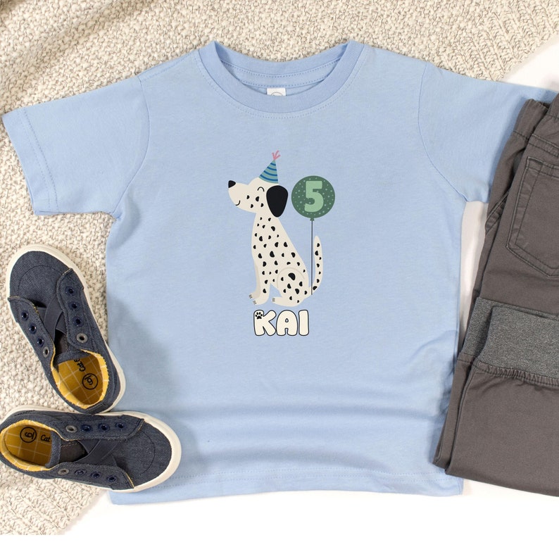 May include: A light blue t-shirt featuring a Dalmatian dog illustration with black spots, wearing a party hat and holding a number 5 balloon. The name "KAI" is printed below the dog. A pair of blue canvas shoes and gray pants are next to the shirt.