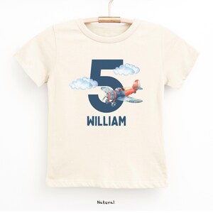 May include: A natural-colored t-shirt featuring a navy blue number 5 with a watercolor airplane and clouds. The name "WILLIAM" is printed below the number in navy blue. The shirt is hanging on a wooden hanger.