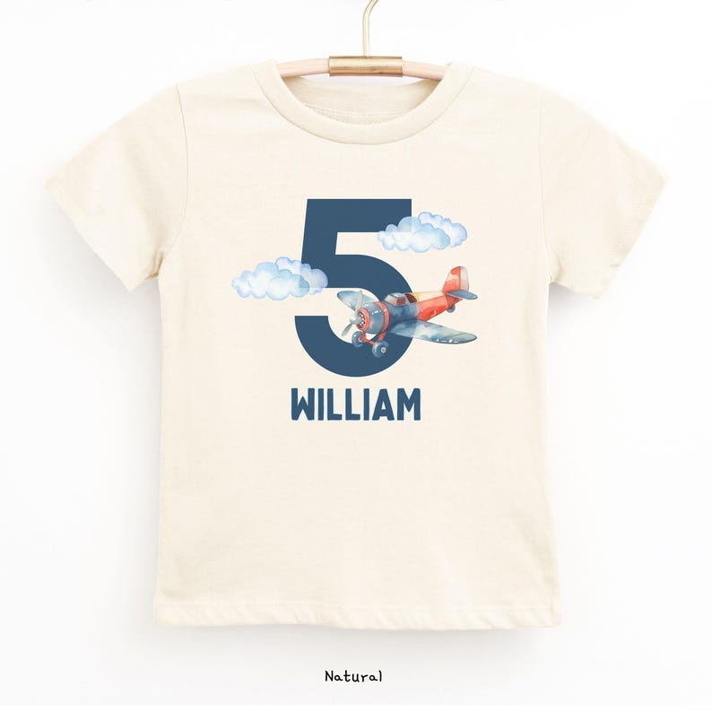 May include: A natural-colored t-shirt featuring a navy blue number 5 with a watercolor airplane and clouds. The name "WILLIAM" is printed below the number in navy blue. The shirt is hanging on a wooden hanger.