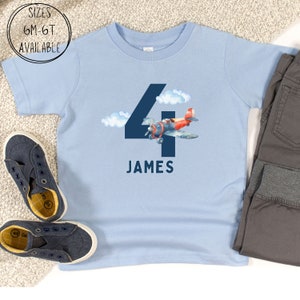 May include: A light blue t-shirt featuring a navy blue number 4 with a watercolor airplane design and the name "JAMES". The shirt also has the text "SIZES 6M-6T AVAILABLE" in a circle. A pair of navy blue shoes and gray pants are next to the shirt.