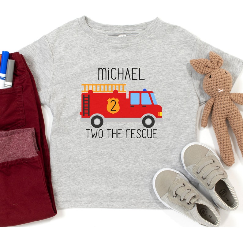 Puede incluir: Una camiseta gris jaspeado de manga corta con un gr&aacute;fico de un cami&oacute;n de bomberos rojo, una escalera amarilla y un escudo con el n&uacute;mero 2. El texto "MICHAEL" aparece sobre el cami&oacute;n y "TWO THE RESCUE" est&aacute; impreso debajo del mismo.