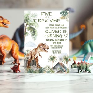 Pode incluir: Um convite de anivers&aacute;rio com tema de dinossauro com o texto "FIVE is a T-Rex VIBE" e "OLIVER IS TURNING 5!". O convite apresenta uma ilustra&ccedil;&atilde;o em aquarela de um T-Rex, palmeiras e um vulc&atilde;o. Dinossauros de brinquedo cercam o convite.