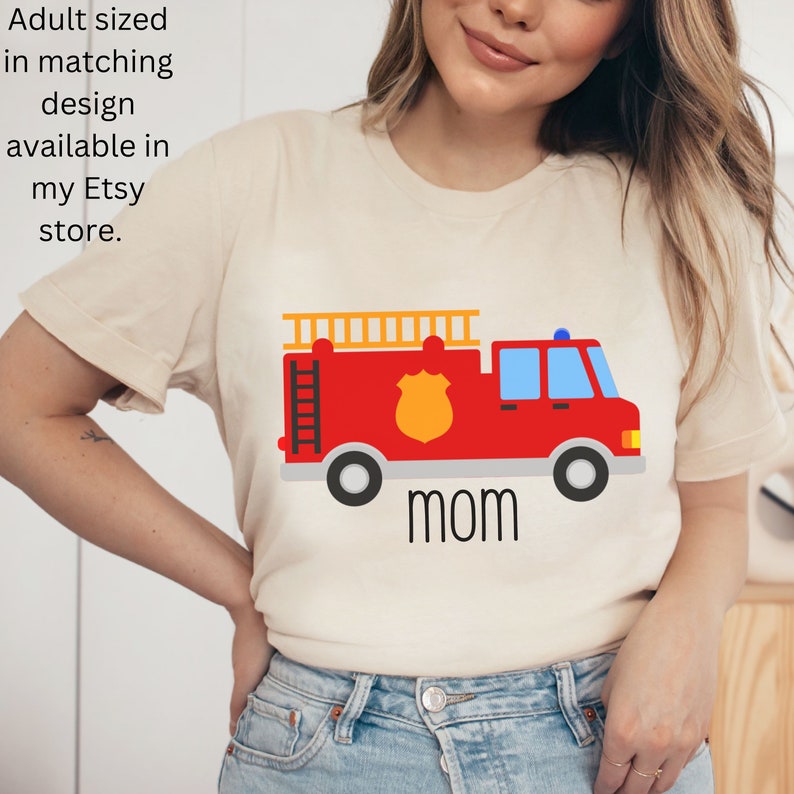 Puede incluir: Una camiseta beige de manga corta con un gr&aacute;fico de un cami&oacute;n de bomberos rojo con una insignia amarilla, una escalera en la parte superior y la palabra mom impresa en negro debajo. El texto Adult sized in matching design available in my Etsy store es visible.