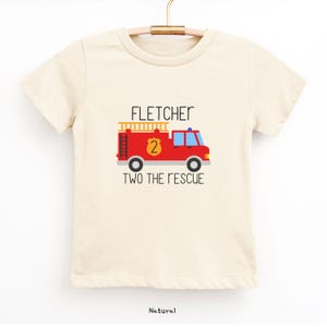 Puede incluir: Una camiseta de manga corta de color natural con un gr&aacute;fico de un cami&oacute;n de bomberos rojo, una escalera amarilla y una insignia con el n&uacute;mero 2. El texto FLETCHER est&aacute; impreso arriba y TWO THE RESCUE est&aacute; impreso debajo del cami&oacute;n.
