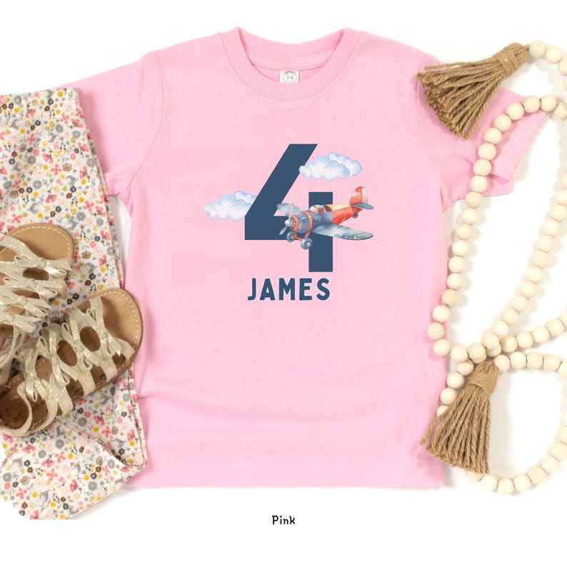 May include: Pink short-sleeved t-shirt with a navy blue number 4 and a watercolor airplane graphic. The name "JAMES" is printed below the number. Floral print pants and gold sandals are visible.