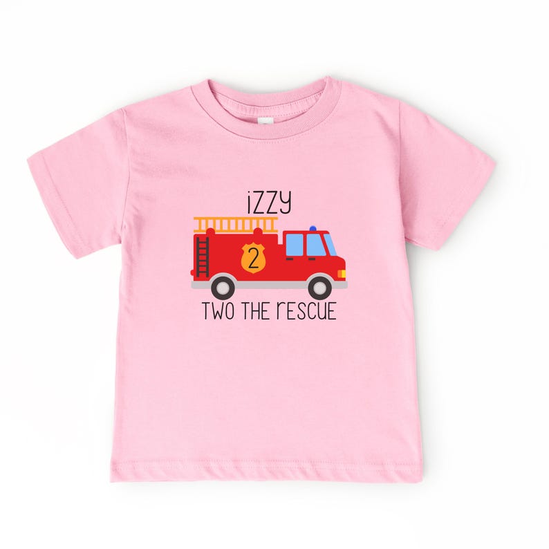 Puede incluir: Una camiseta de manga corta de color rosa claro con el gr&aacute;fico de un cami&oacute;n de bomberos rojo, una escalera amarilla y un escudo con el n&uacute;mero 2. El texto "iZZy" aparece sobre el cami&oacute;n y "TWO THE RESCUE" aparece debajo.