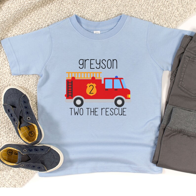 Puede incluir: Una camiseta de manga corta de color azul claro con un gr&aacute;fico de un cami&oacute;n de bomberos rojo con una escalera amarilla, un escudo amarillo con el n&uacute;mero 2 y el texto "greyson" arriba y "TWO THE RESCUE" abajo en letras negras.