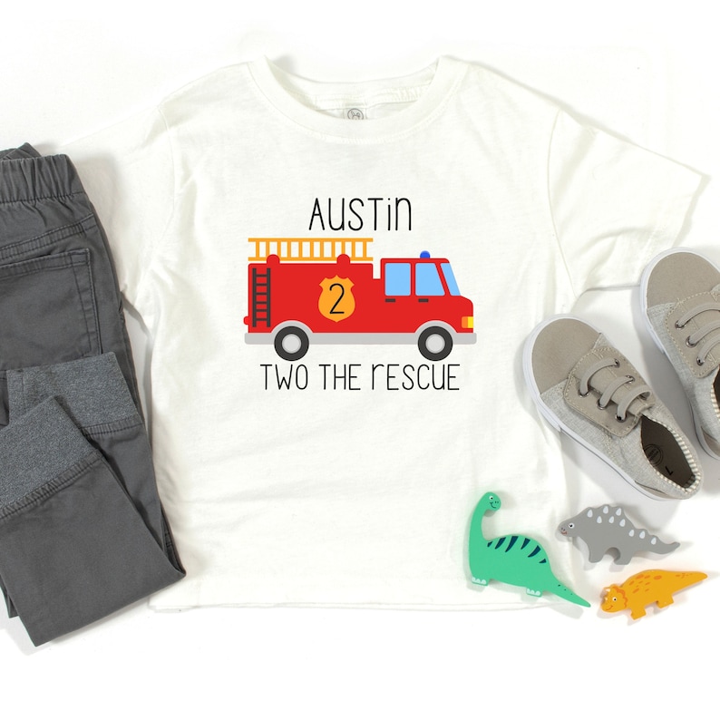 Puede incluir: Una camiseta blanca con un gr&aacute;fico de un cami&oacute;n de bomberos rojo, un escudo amarillo con el n&uacute;mero 2, el nombre AUSTIN y el texto TWO THE RESCUE. La camiseta se muestra con pantalones de ch&aacute;ndal grises, zapatillas grises y figuras de dinosaurios.