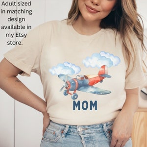 May include: Beige t-shirt featuring a watercolor illustration of a red, blue, and yellow airplane with blue clouds. The word "MOM" is printed below the plane. The text "Adult sized in matching design available in my Etsy store" is also visible.