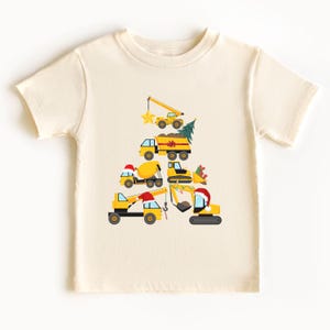 May include: A cream-colored short-sleeve t-shirt featuring a graphic of yellow construction vehicles stacked in the shape of a Christmas tree. The design includes a crane, dump truck, cement mixer, and excavator adorned with Santa hats and a star.