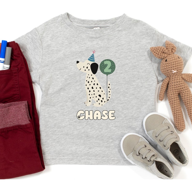 May include: A light gray t-shirt featuring a Dalmatian dog wearing a party hat and holding a number 2 balloon. The name "CHASE" is printed below the dog. The shirt is paired with red pants, gray shoes, and a stuffed bunny.