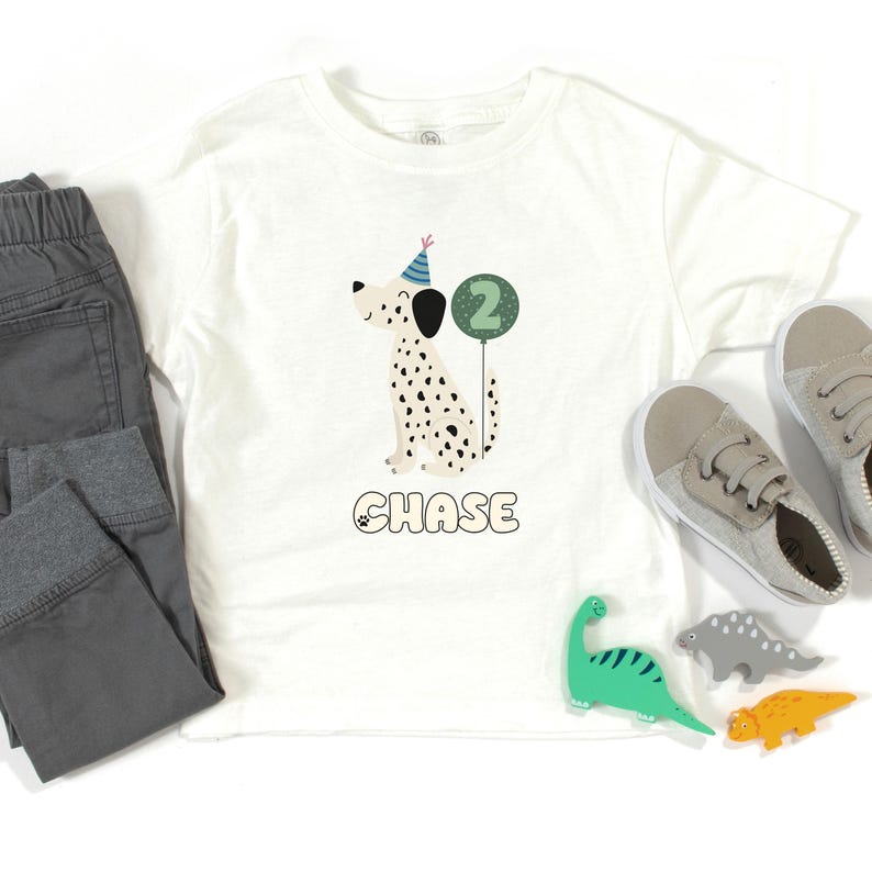 May include: White t-shirt featuring a cartoon Dalmatian wearing a party hat and holding a number 2 balloon. The name "CHASE" is printed below the dog. Gray pants and shoes are next to the shirt, along with dinosaur toys.