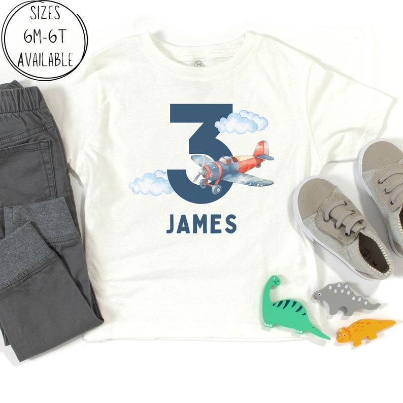 May include: White t-shirt with a navy blue number 3, an airplane graphic, and the name "JAMES" in navy blue. The shirt is paired with gray pants and sneakers. Sizes 6M-6T available.