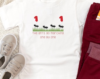 May include: A white short-sleeved t-shirt featuring a graphic of four black ants walking on green grass, with two red balloons shaped like the number one attached to the ants. The text reads, The ants go marching one by one in red letters.