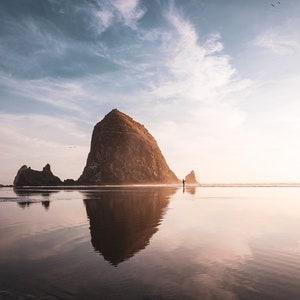 May include: A large rock formation, known as Haystack Rock, rises from the sandy beach. The rock is reflected in the calm, still water. The sky is a soft blue with white clouds.