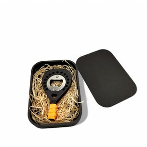 May include: A black paddle-shaped bottle opener with an orange handle, in a black box with shredded paper.