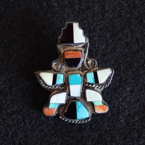 May include: A silver and turquoise pendant featuring a stylized figure with intricate details. The figure is adorned with black, white, and orange stones.