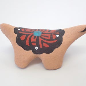 May include: A small, tan-colored ceramic animal figurine with a decorative saddle. The saddle features a black border with red and white accents, and a central turquoise design. The figurine has a smooth, rounded form and is set against a white background.