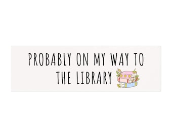 May include: A rectangular sticker with the text "PROBABLY ON MY WAY TO THE LIBRARY" in black, sans-serif font. A stack of books with floral accents is to the right of the text. The books are pink, blue, and yellow.