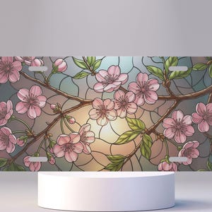 May include: Rectangular license plate featuring a stained-glass design of pink cherry blossoms on brown branches with green leaves. The background has a gradient of blue, gray, and yellow hues. The plate has four mounting holes.