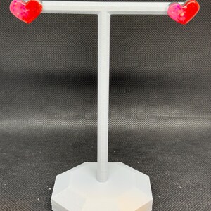 May include: A pair of heart-shaped earrings with pink and red resin. The earrings are displayed on a white jewelry stand.
