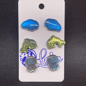 May include: A set of six sparkly stud earrings featuring a manatee, dolphin, and jellyfish design. The earrings are displayed on a white card with the word "Love" written on it.