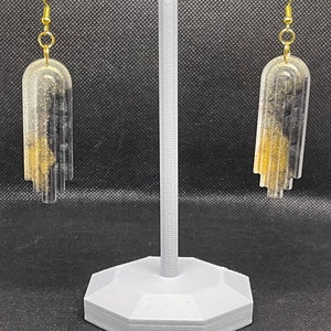 May include: A pair of gold and black resin earrings with a geometric design. The earrings are hanging from a gray earring stand.