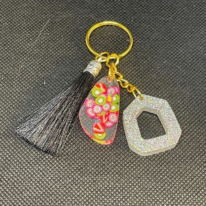 May include: A gold key chain with a black tassel, a clear resin charm with a pink and green fruit design, and a silver glitter charm in the shape of a hexagon.