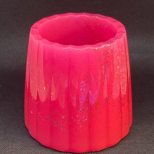 May include: A pink, ribbed, cylindrical vase with iridescent glitter embedded in the resin. The vase is made of resin and has a smooth, glossy finish.