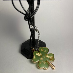 May include: A four-leaf clover pendant necklace. The pendant is light green with gold leaf accents and a gold-colored border. The necklace has a black cord and a silver-colored chain.