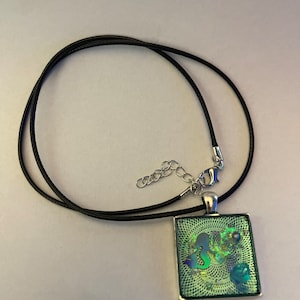 May include: A silver pendant necklace with a square green and blue iridescent glass pendant featuring a mermaid silhouette. The pendant is on a black cord.