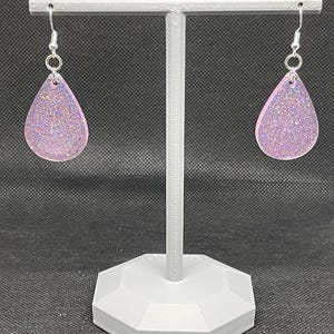 May include: A pair of teardrop-shaped earrings with a light pink glitter finish. The earrings are hanging from silver hooks on a white display stand.