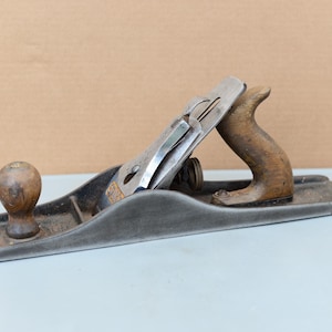 May include: A vintage hand plane, featuring a wooden handle and knob, with a steel blade. The plane has a gray body with signs of wear, indicating its age and use. The wooden components show a natural, aged finish.