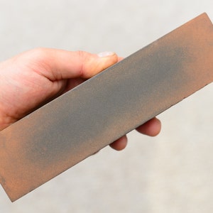 May include: A rectangular sharpening stone with a brown and gray surface. The stone is held in a hand.