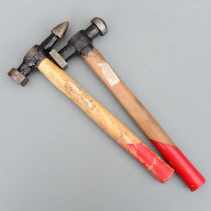 Puede incluir: Dos martillos de metal con mangos de madera. Uno tiene una cabeza puntiaguda, el otro una cabeza redonda. Los mangos son de color marrón claro con extremos pintados de rojo. Los martillos están sobre una superficie gris.
