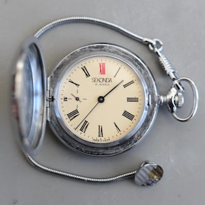 Może przedstawiać: Srebrny zegarek kieszonkowy Sekonda z kremową tarczą i czarnymi rzymskimi cyframi. Zegarek posiada łańcuszek i zawiasową pokrywę. Cyfra XII jest czerwona. Na tarczy widnieje napis "Sekonda 16 Jewels" i "Made in USSR".