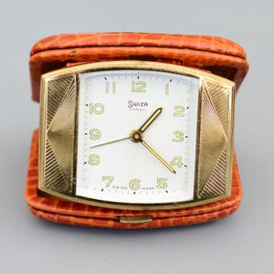 May include: A vintage Swiza Coral travel clock with a gold-tone rectangular face and a white dial. The clock has gold hands and numerals, and is encased in an orange leatherette case. The clock is marked "SWISS MADE".