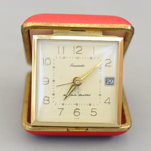 Pode incluir: Um despertador Timemaster vintage em uma caixa vermelha e dourada. O mostrador quadrado do relógio tem ponteiros e algarismos dourados, com um visor de data. O relógio está aberto, revelando o mostrador e a moldura dourada.