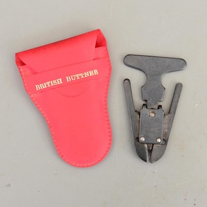 May include: A vintage metal tool with a T-shaped handle and a cutting edge, alongside a red leather case. The case is labelled "BRITISH BUTTNER" in gold lettering. The tool appears to be a specialized cutting instrument.
