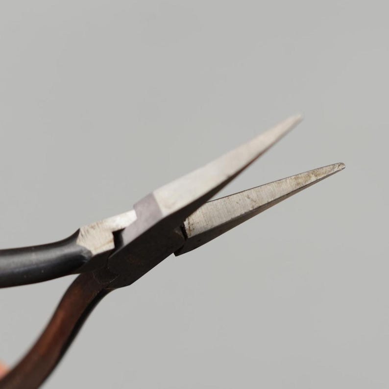 May include: Close-up of needle-nose pliers with a black handle and silver-colored jaws. The pliers are open, revealing the pointed tips. The tool is designed for precision work, commonly used in jewelry making and electronics.