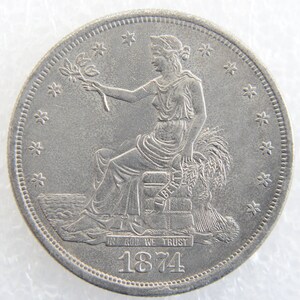 Puede incluir: Una moneda de plata de un dólar con una figura de la Libertad sentada sosteniendo una rama de olivo y una gavilla de trigo. La moneda tiene la inscripción "In God We Trust" y la fecha "1874".