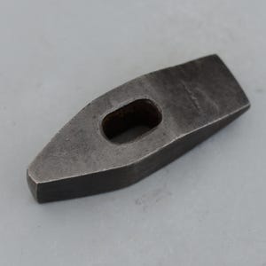May include: A dark gray, heavy-duty metal hammer head with a rectangular body and a tapered, pointed end. The hammer head features a central, oval-shaped hole for the handle. The tool has a worn, industrial appearance.