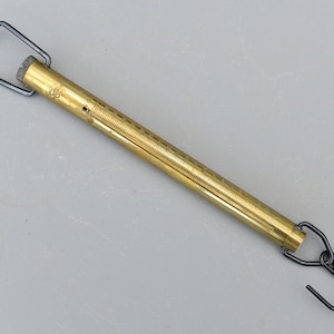May include: A brass spring scale with black metal hooks at each end. The scale has a graduated scale on the side.
